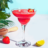 A chilled strawberry margarita in a stemmed glass, garnished with a slice of strawberry, sits on a white table next to a whole strawberry, a lime wedge, and a wooden citrus reamer, with tropical leaves in the background.