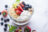 A clear glass bowl filled with yogurt, granola, sliced bananas, strawberries, blueberries, and blackberries, set on a white marble surface with scattered fresh berries and mint leaves.